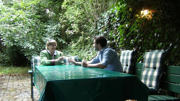 Ein gemütlicher Garten mit einem Tisch und zwei Personen, die sich unterhalten. Die Umgebung ist grün und von Pflanzen umgeben.