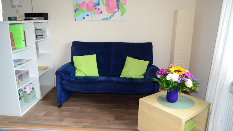 Ein gemütlicher Wohnraum mit einem blauen Sofa und hellgrünen Kissen. Der Tisch hat eine Blumenvase und im Hintergrund steht ein Bücherregal.