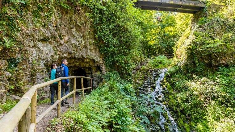 Zwei Personen stehen vor einer Höhle in einer grünen, bewaldeten Umgebung mit einem kleinen Wasserfall und einer Brücke darüber.