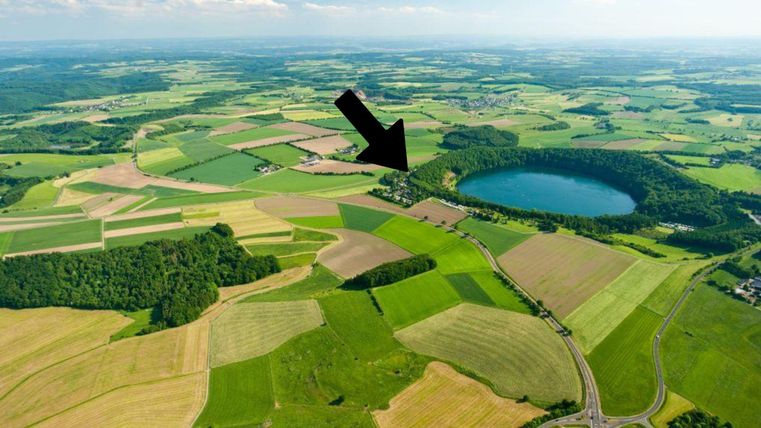 Eine weite, grüne Landschaft mit Feldern und Wäldern. Im Hintergrund befindet sich ein klarer Teich.