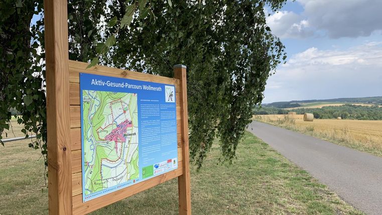 Ein Informationsschild steht an einem Weg in der Natur. Im Hintergrund sind Felder und eine hügelige Landschaft zu sehen.