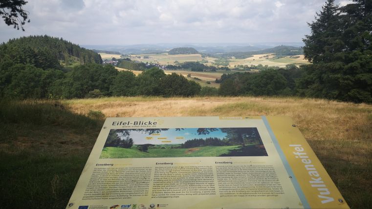 Eine Aussichtstafel mit Informationen über die Eifel. Im Hintergrund erstrecken sich grüne Hügel und ein bewölter Himmel.