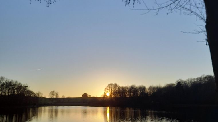Ein ruhiger See unter einem klaren Himmel bei Sonnenuntergang. Die Spiegelung des Lichts auf dem Wasser schafft eine friedliche Atmosphäre.