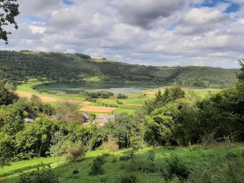 Eine beeindruckende Landschaft mit einem ruhigen See in der Mitte. Umgeben von grünen Wiesen und sanften Hügeln unter einem bewölkten Himmel.
