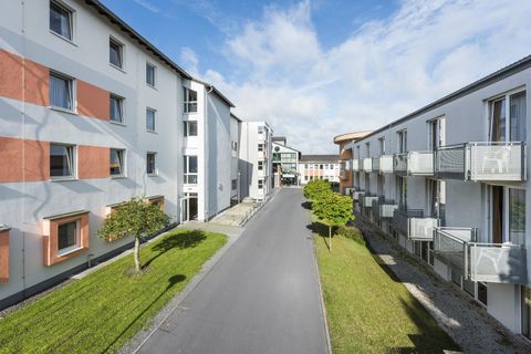Eine ruhige Wohnstraße zwischen modernen Mehrfamilienhäusern. Im Hintergrund sind ein paar Bäume und ein klarer Himmel zu sehen.