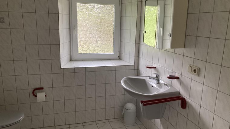 Ein einfaches Badezimmer mit weißen Fliesen und roten Akzenten. Es gibt ein Fenster, ein Waschbecken und eine Toilette.