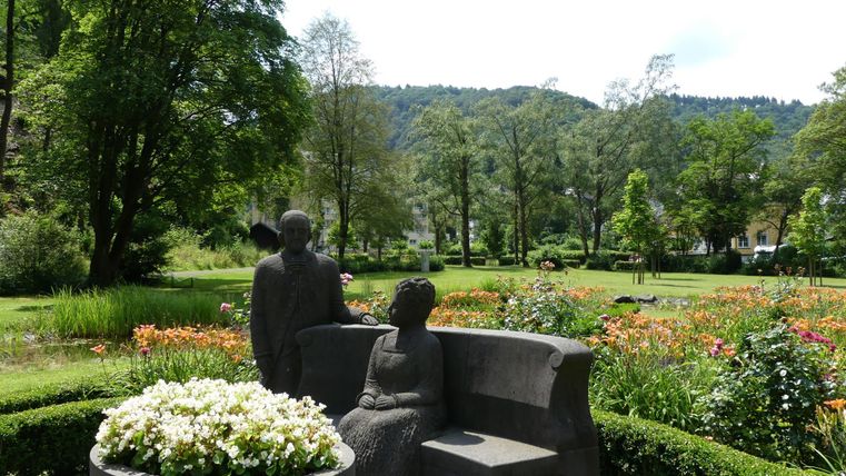 Eine ruhige Parklandschaft mit bunten Blumen und Bäumen. Im Vordergrund stehen zwei Sitzfiguren auf einer Bank.