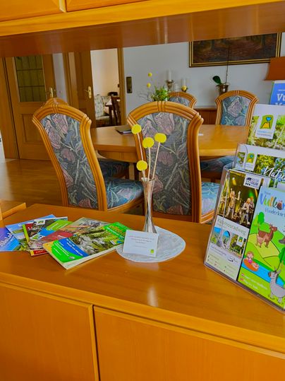 Ein einladender Raum mit einem Holzschreibtisch und einem großen Esstisch im Hintergrund. Auf dem Schreibtisch liegen Informationsbroschüren und eine kleine Vase mit gelben Blumen.