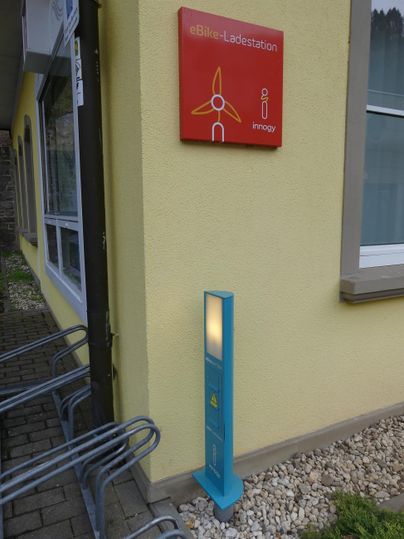 Ein Informationsschild an einer gelben Wand, neben einem blauen Stehen. Der Bereich ist mit Kieselsteinen gestaltet und es stehen einige Fahrradständer in der Nähe.