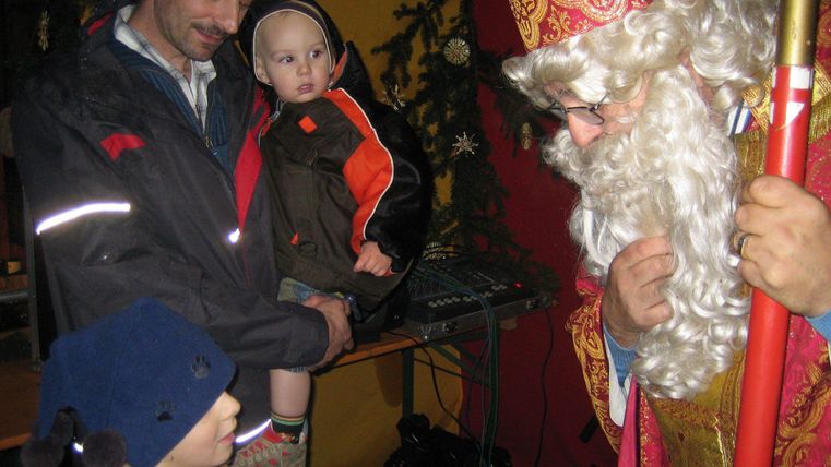Ein Mann steht mit zwei Kindern, während ein freundlicher Weihnachtsskulptur ihnen gegenübersteht. Die Umgebung zeigt eine festliche Atmosphäre.