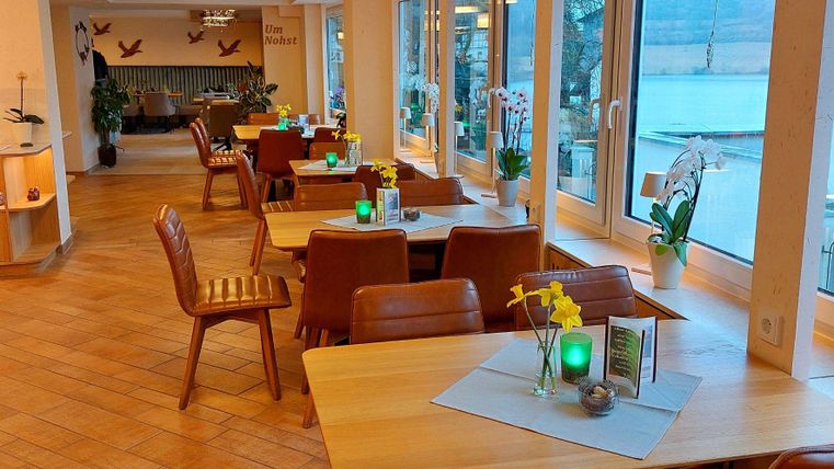 Ein gemütliches Restaurant mit Holztischen und Stühlen. Große Fenster bieten einen Blick auf das Wasser und sorgen für eine helle Atmosphäre.