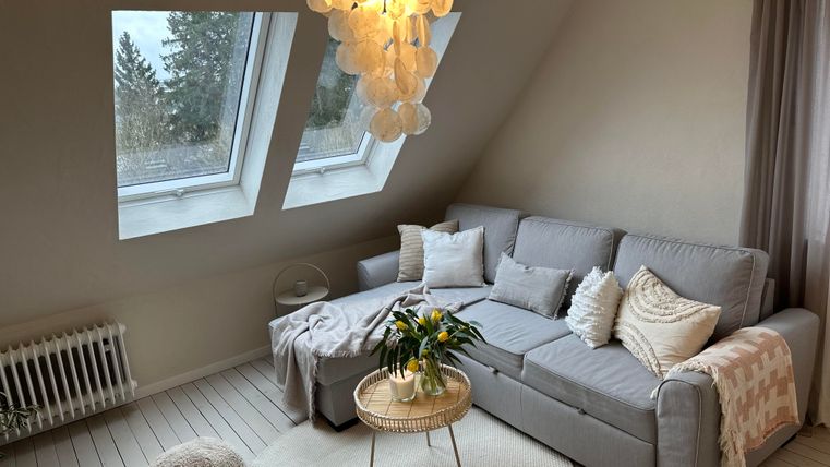 Ein gemütliches Wohnzimmer mit einem grauen Sofa und einer runden Kaffeetisch. Große Fenster lassen viel Tageslicht herein und die Dekoration ist modern und einladend.