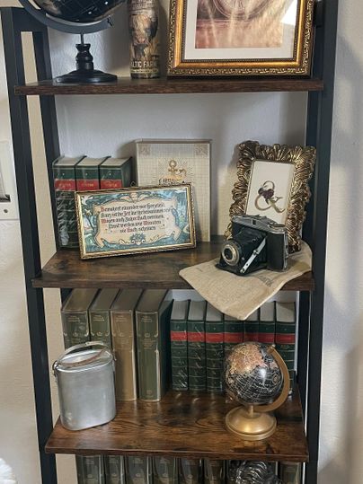 Ein Bücherregal mit alten Büchern, dekorativen Gegenständen und einer Weltkugel. Darunter sind ein gerahmtes Bild und eine alte Kamera zu sehen.