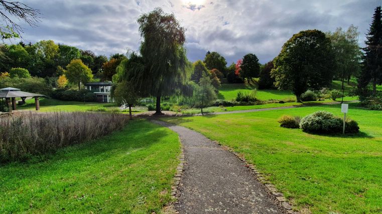 Ein malerischer Weg führt durch einen gepflegten Park mit üppigem Grün. Im Hintergrund sind Bäume und ein bewölkter Himmel sichtbar.