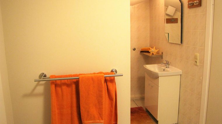 Ein helles Badezimmer mit weißen Wänden und Fliesen. An der Wand hängt ein orangefarbenes Handtuch, und es gibt einen kleinen Waschplatz mit Spiegel.