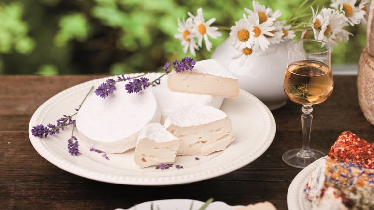 Eine schöne Anricht platter mit verschiedenen Käsesorten, dekoriert mit Lavendel. Im Hintergrund sind frische Blumen und ein Glas Wein zu sehen.