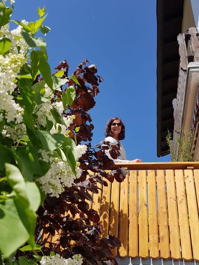 Eine Person steht auf einem Balkon und schaut lächelnd in die Kamera. Im Vordergrund sind blühende Pflanzen zu sehen, und der Himmel ist klar und blau.