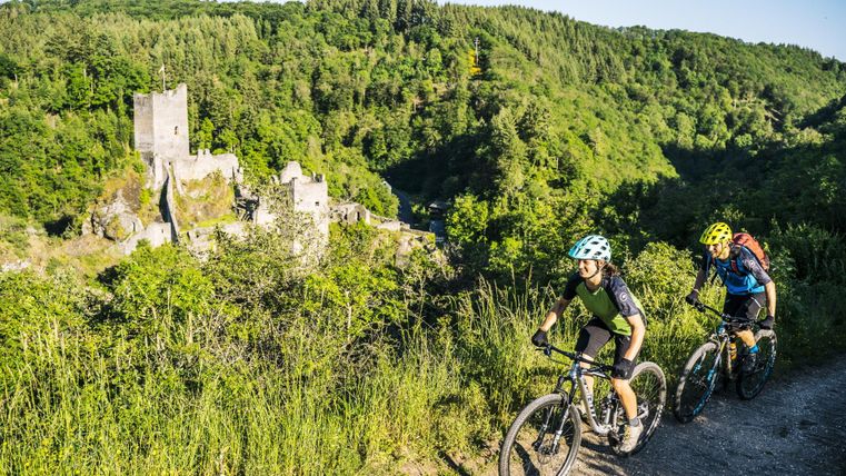 Zwei Mountainbiker fahren auf einem Weg durch üppige grüne Landschaft. Im Hintergrund ist eine alte Burgruine sichtbar.