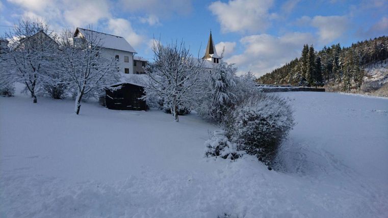 Eine verschneite Landschaft mit einem weißen Feld und schneebedeckten Bäumen. Im Hintergrund sind ein Haus und ein Kirchturm zu sehen.
