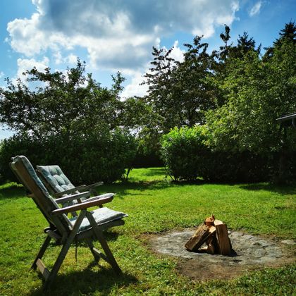 Ein gemütlicher Garten mit einem Stuhl und einem kleinen Lagerfeuer. Umgeben von Bäumen und grünem Gras unter einem blauen Himmel.