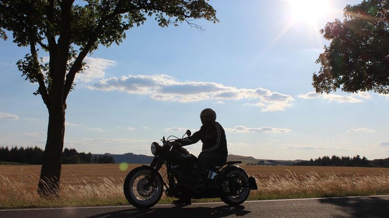Ein Motorradfahrer fährt auf einer Landstraße, umgeben von Bäumen und einem weiten Feld. Die Sonne scheint hell am Himmel und schafft eine entspannte Atmosphäre.