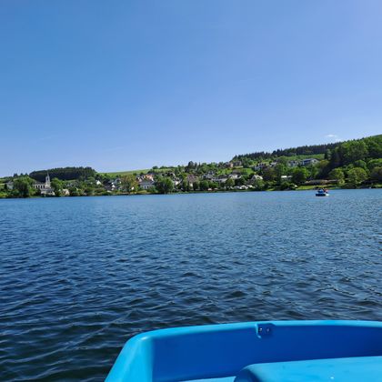 Ein ruhiger See mit klarem Wasser und einer schönen Uferlandschaft. Im Hintergrund sind grüne Hügel und einige Gebäude zu sehen.
