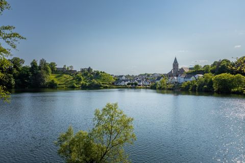Ein ruhiger See umgeben von grünen Hügeln und Bäumen. Im Hintergrund sind Häuser und eine Kirche zu sehen.