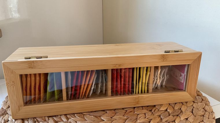 Eine elegante Holzkiste mit transparentem Fenster, die verschiedene Teebeutel in leuchtenden Farben enthält. Die Kiste steht auf einem geflochtenen Untergrund.