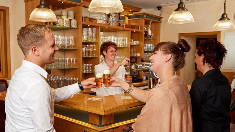 Eine fröhliche Bartenderszene in einer gemütlichen Bar. Zwei Gäste stoßen mit ihren Bieren an, während die Barkeeperin lächelt.