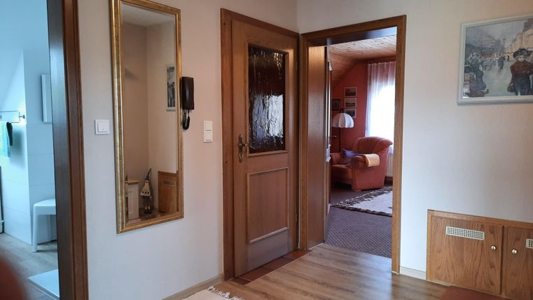 Ein heller Flurbereich mit einem Spiegel und einer Holzinnentür. Im Hintergrund ist ein gemütliches Wohnzimmer mit einem roten Sofa sichtbar.