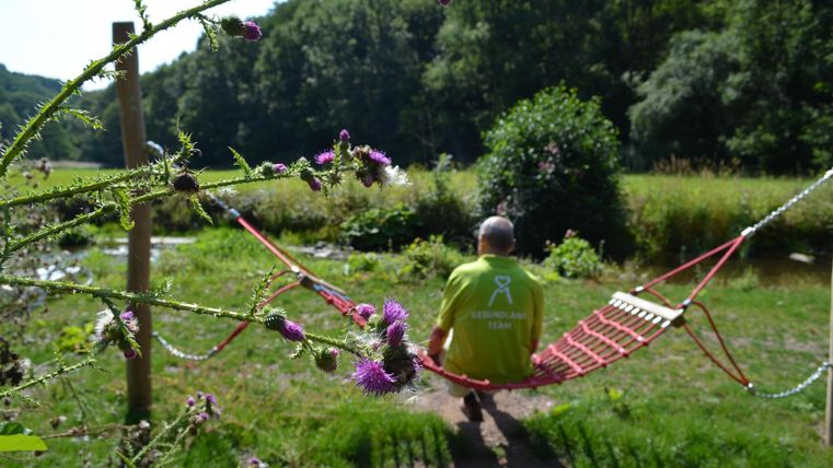 Eine Person sitzt in einer Hängematte und genießt die ruhige Natur am Ufer eines Flusses. Im Hintergrund sind Bäume und Blumen sichtbar.