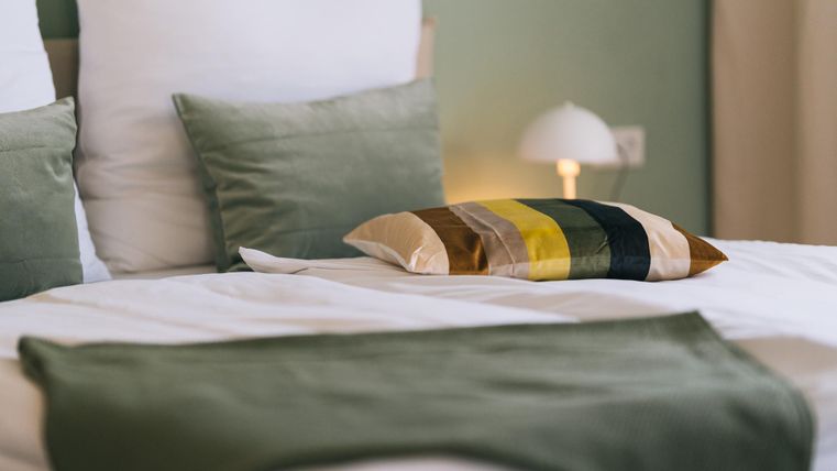 Ein gemütliches Bett mit grünen und bunten Kissen. Die ruhige Farbpalette schafft eine entspannte Atmosphäre.