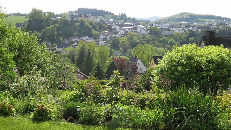Eine malerische Landschaft mit einem schönen Garten im Vordergrund und einer kleinen Stadt im Hintergrund. Die Umgebung ist grün und hügelig, unter einem hellen Himmel.