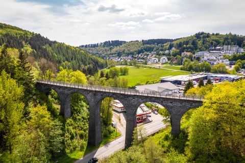Eine beeindruckende Steinbrücke erhebt sich über eine grüne Landschaft. Umgeben von Wäldern und kleinen Gebäuden verleiht sie dem Bild eine charmante Atmosphäre.