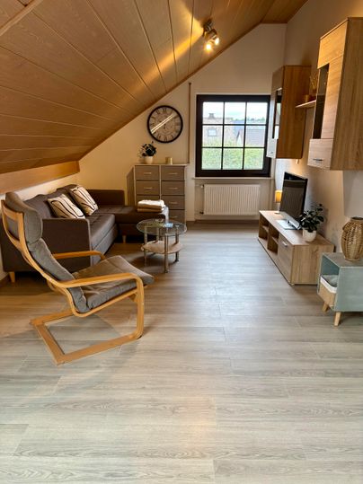 A cozy living room in the attic with a sofa and an armchair. There is a table, a TV corner, and large windows with a view outside.