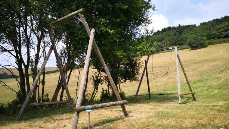 Ein Spielplatz mit zwei Schaukeln aus Holz in einer grünen Wiese. Im Hintergrund sind Bäume und sanfte Hügel zu sehen.