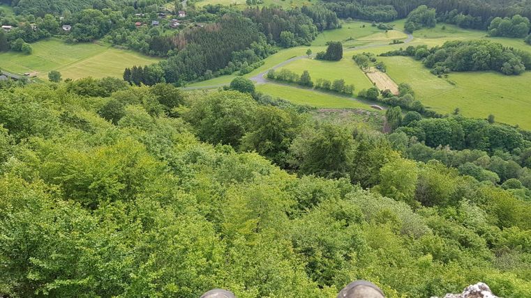 Eine beeindruckende Aussicht auf eine grüne Landschaft mit Wäldern und Wiesen. Im Vordergrund sind Stiefel auf einem Felsen sichtbar.