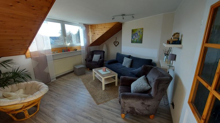 Ein gemütliches Wohnzimmer mit einer Sitzecke und einem hellen Fenster. Die Einrichtung ist modern und einladend, mit einem Teppich und Pflanzen.