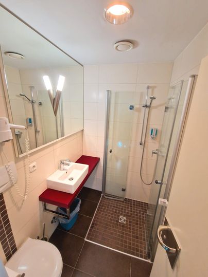 Ein modernes Badezimmer mit einer Duschkabine und einem Waschbecken auf einer roten Ablage. Die Wände sind hell gefliest und der Boden ist dunkel gefliest.