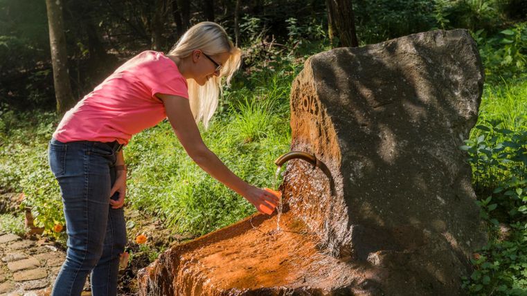 Eine Frau mit blonder Haaren füllt ein Glas an einem natürlichen Wasserlauf. Im Hintergrund sind Bäume und grüne Pflanzen zu sehen.