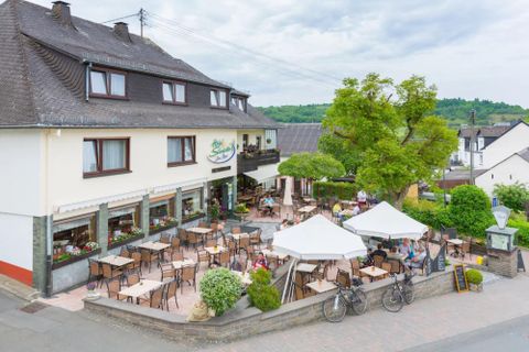 A cozy café with a terrace and many outdoor tables. In the background, trees and a peaceful environment can be seen.