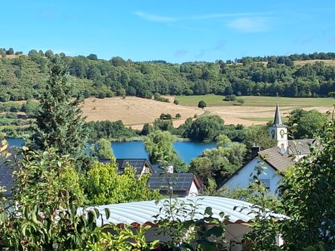Ein malerischer Blick auf eine grüne Landschaft mit Feldern und einem Gewässer. Im Hintergrund sind ein Dorf und eine Kirche zu sehen.