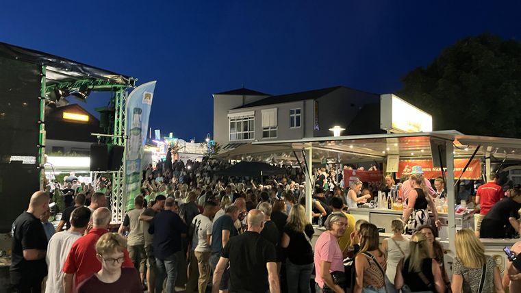 Eine große Menschenmenge auf einem Festplatz bei Nacht. Stände mit Essen und Getränken sind im Vordergrund zu sehen.