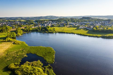 Eine malerische Landschaft mit einem ruhigen See und grünen Wiesen. Im Hintergrund sind kleine Häuser und Hügel sichtbar.