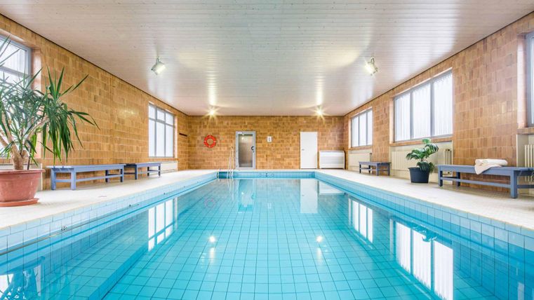 Ein helles Hallenbad mit klarem Wasser und großen Fenstern. Pflanzen und Sitzgelegenheiten ergänzen den Raum.