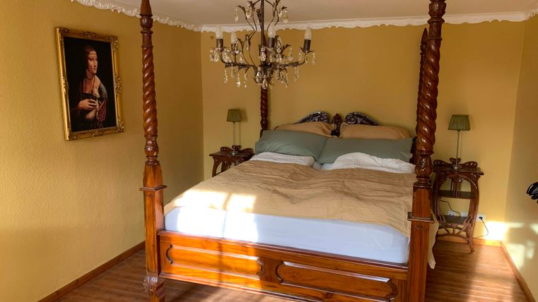 Ein gemütliches Schlafzimmer mit einem Himmelbett und einem eleganten Kronleuchter. An der Wand hängt ein klassisches Gemälde.