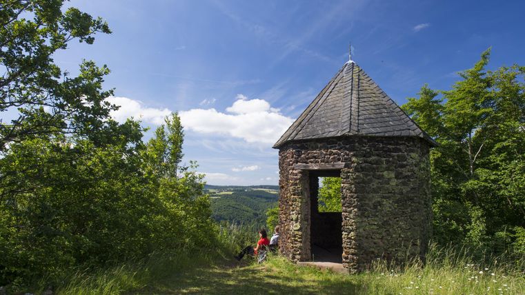 Ein historischer Aussichtspunkt aus Stein in einer grünen Umgebung. Im Vordergrund entspannen sich Personen im grasbewachsenen Gelände.
