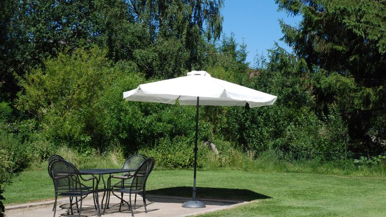 Ein ruhiger Garten mit einem Tisch und Stühlen unter einem weißen Sonnenschirm. Umgeben von üppigem Grün und Bäumen.