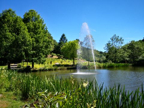 Ein Teich im Kurpark mit einer Wasserfontäne, umgeben von grünen Bäumen und Pflanzen unter klarem blauem Himmel.
