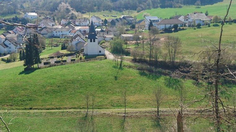Eine malerische Landschaft mit einem kleinen Dorf und einer Kirche. Umgeben von grünen Wiesen und sanften Hügeln.